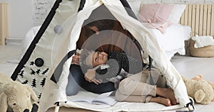 Mother and son are reading a book in tepee together, boy laying on woman`s legs.