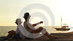 Mother and son sit on the beach at sunset watching a ship go by