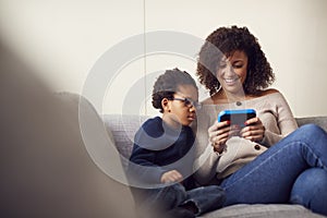 Mother And Son Sitting On Sofa At Home Playing Computer Game Together On Hand Held Device At Home