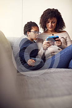 Mother And Son Sitting On Sofa At Home Playing Computer Game Together On Hand Held Device At Home