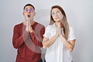 Mother and son standing together over isolated background begging and praying with hands together with hope expression on face