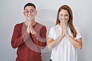 Mother and son standing together over isolated background praying with hands together asking for forgiveness smiling confident