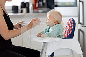 Mother spoon feeding her baby boy child in baby chair with fruit puree in kitchen at home. Baby solid food introduction