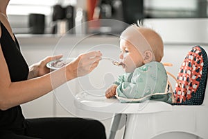 Mother spoon feeding her baby boy child in baby chair with fruit puree in kitchen at home. Baby solid food introduction