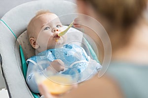 Mother spoon feeding her baby boy infant child in baby chair with fruit puree. Baby solid food introduction concept.