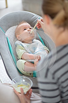 Mother spoon feeding her baby boy infant child in baby chair with fruit puree. Baby solid food introduction concept.