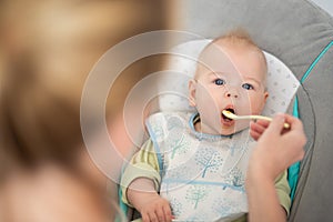 Mother spoon feeding her baby boy infant child in baby chair with fruit puree. Baby solid food introduction concept.