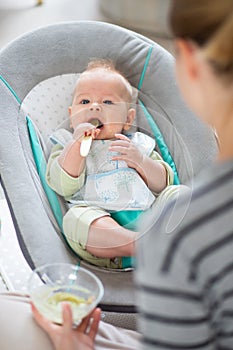 Mother spoon feeding her baby boy infant child in baby chair with fruit puree. Baby solid food introduction concept.