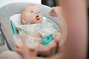 Mother spoon feeding her baby boy infant child in baby chair with fruit puree. Baby solid food introduction concept.