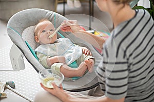 Mother spoon feeding her baby boy infant child in baby chair with fruit puree. Baby solid food introduction concept.