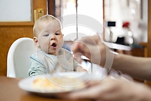 Mother spoon feeding her infant baby boy child sitting in high chair at the dining table in kitchen at home