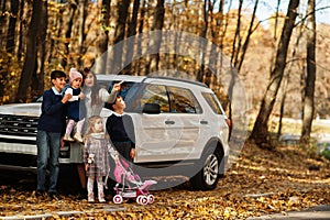 Mother stand near white suv car with four kids in autumn park. Family walk in fall forest