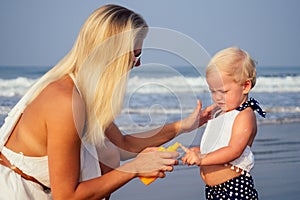 Mother in stylish glasses and white dress is putting orange spray bottle SPF on newborn baby one year old daughter a
