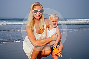 Mother in stylish glasses and white dress is putting orange spray bottle SPF on newborn baby one year old daughter a