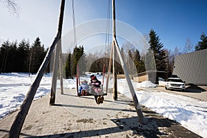Mother swinging with daughters in big wooden swing in early spring snowy mountains against tiny house and car
