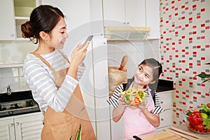 Mother takes a photo of vegetable salad with a little girl in a kitchen at home. Leisure activity