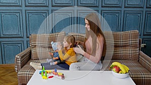 Mother taking care, brushing hair of daughter child girl in living room, kid playing with toy doll