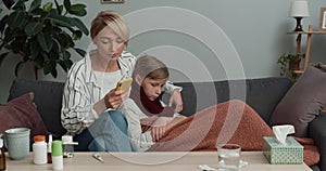 Mother taking and looking at thermometer while sitting on sofa with ill kid. Worried woman calling doctor on smartphone