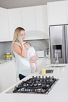 Mother talking on mobile phone while carrying her baby in kitchen