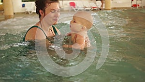 Mother teaches her baby to swim in a closed public pool. The child is swinging on an artificial wave in the aquapark