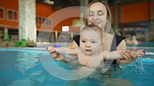 Mother teaches her baby to swim in a closed public pool. The child is swinging on an artificial wave in the aquapark