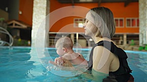 Mother teaches her baby to swim in a closed public pool. The child is swinging on an artificial wave in the aquapark