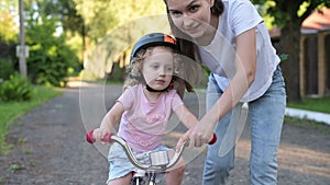mother teaches her daughter to ride a bike. Little girl in helmet learns to ride