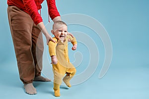 Mother teaches toddler baby to stand on his feet and walk, child boy on studio blue background