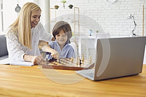 Mother teaching son to play chess following online lesson on laptop at home. Parent and child playing board game