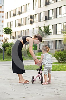 Mother teaching toddler girl to ride on first bike without pedals on road at town. Summer day