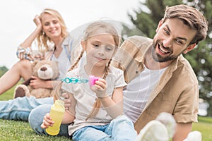mother with teddy bear looking at happy father and daughter blowing soap bubbles
