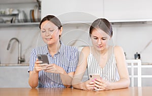 Mother and teen daughter looking at their phones sitting at kitchen-table
