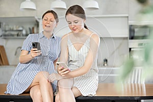 Mother and teen daughter looking at their phones sitting on kitchen-table