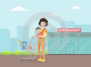Mother with Toddler Baby in Sling Pushing Shopping Cart, Young Woman With Her Kid Standing in Front of Supermarket