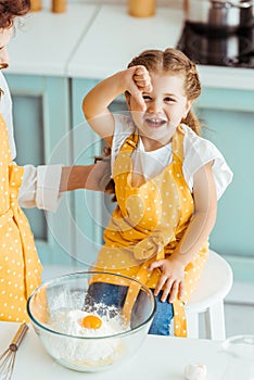 Mother touching daughter in apron near bowl with flour and smashed egg while excited child