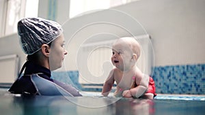 Mother or trainer explain to little baby boy who is sitting on the side of indoors swimming pool what she will do. Push