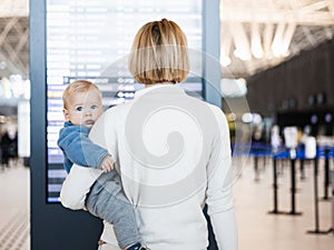 Mother traveling with child, holding his infant baby boy at airport terminal, checking flight schedule, waiting to board