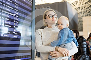 Mother traveling with child, holding his infant baby boy at airport terminal, checking flight schedule, waiting to board