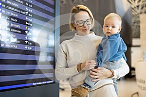 Mother traveling with child, holding his infant baby boy at airport terminal, checking flight schedule, waiting to board