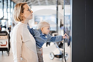 Mother traveling with child, holding his infant baby boy at airport terminal, checking flight schedule, waiting to board