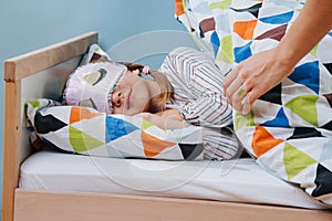 Mother tucks her little daughter in bed. Girl wearing sleeping mask