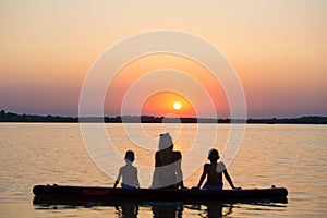 Mother and two children sit on on paddle board and look at the sunset.Water sports , active lifestyle.