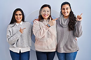 Mother and two daughters standing over blue background cheerful with a smile on face pointing with hand and finger up to the side