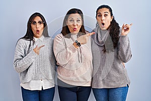 Mother and two daughters standing over blue background surprised pointing with finger to the side, open mouth amazed expression