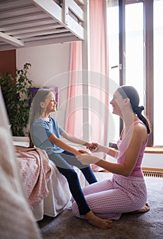 Mother waking daughter up for school in morning. She kneeling by child& x27;s bed and holding daughter by hands.