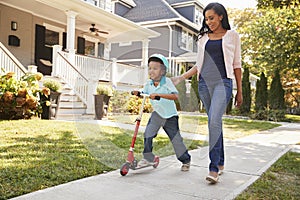 Mother Walks With Son As He Rides Scooter Along Sidewalk