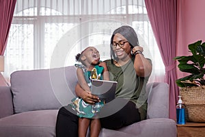 Mother was teaching the daughter through tablet joyfully while sitting on the sofa. black people or African Americans. Home study