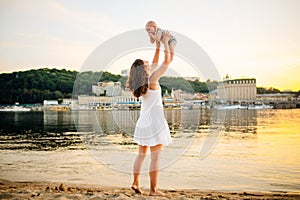 Mother which turns the child against a sunset and water. Happy mom and baby. Playing on beach. Young woman tossing up her son