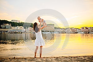 Mother which turns the child against a sunset and water. Happy mom and baby. Playing on beach. Young woman tossing up her son