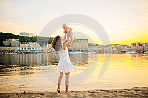 Mother which turns the child against a sunset and water. Happy mom and baby. Playing on beach. Young woman tossing up her son